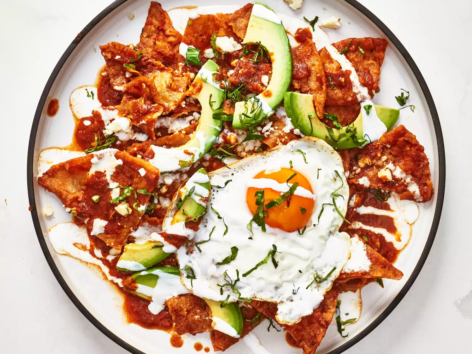 Overhead view of chilaquiles with fried egg and sliced avocado on white background