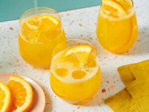 Three glasses with a yellow citrus beverage garnished with orange slices, placed on a terrazzo surface near a napkin and orange slices
