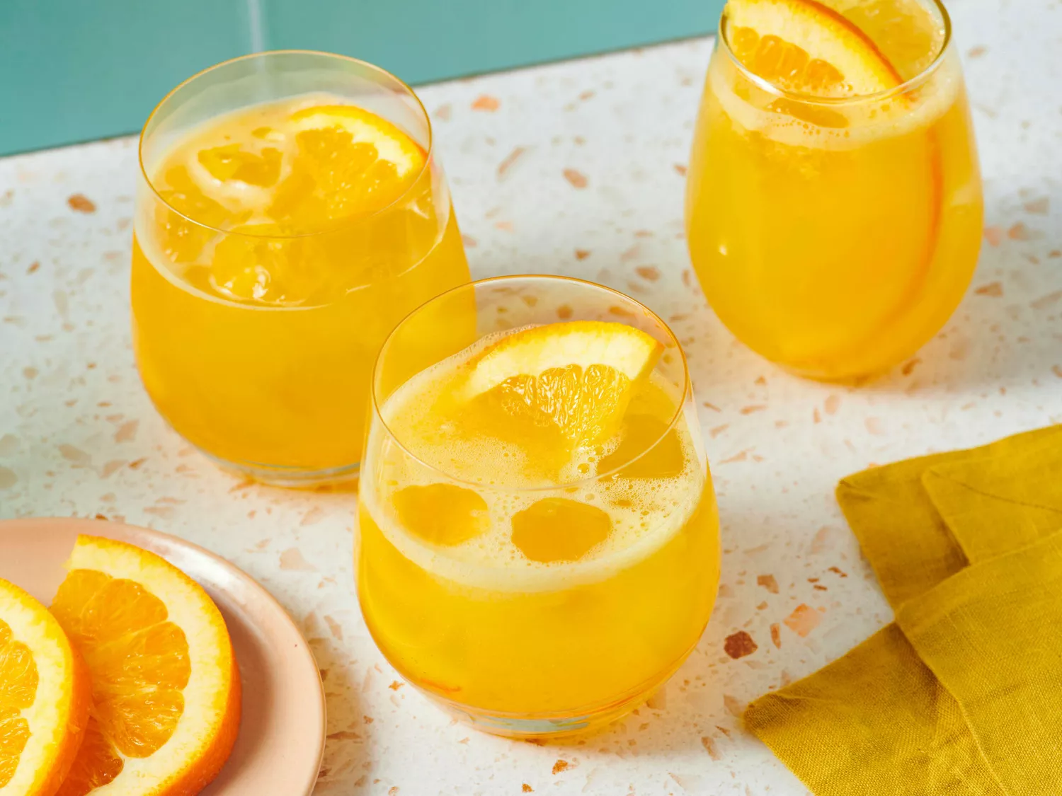 Three glasses with a yellow citrus beverage garnished with orange slices, placed on a terrazzo surface near a napkin and orange slices