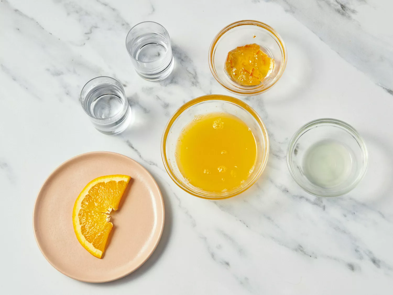 Ingredients for making a breakfast margarita, including orange juice, an orange wedge, and small glasses of liquid on a countertop