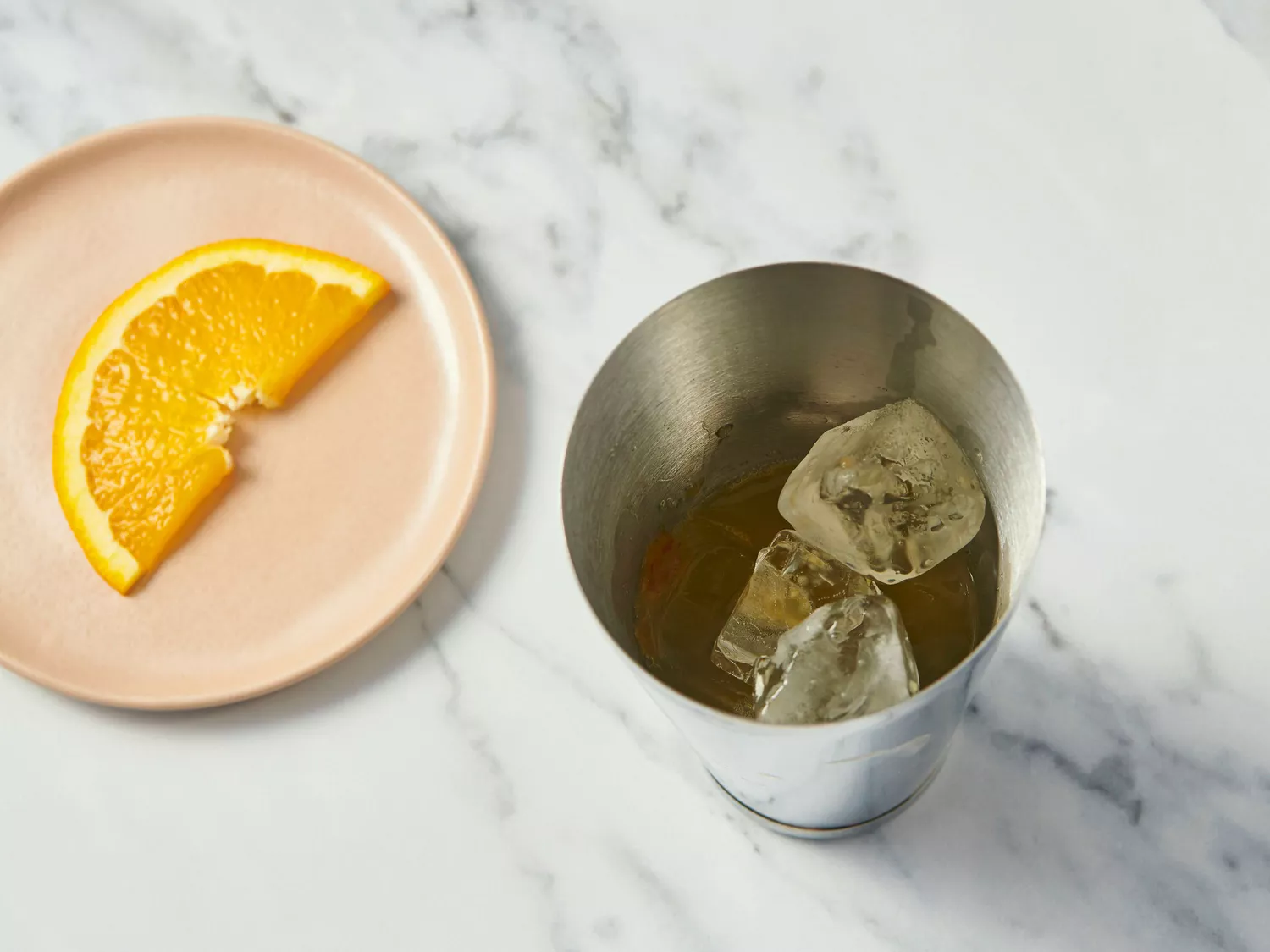 Orange slice on a plate next to a metal cup containing ice cubes