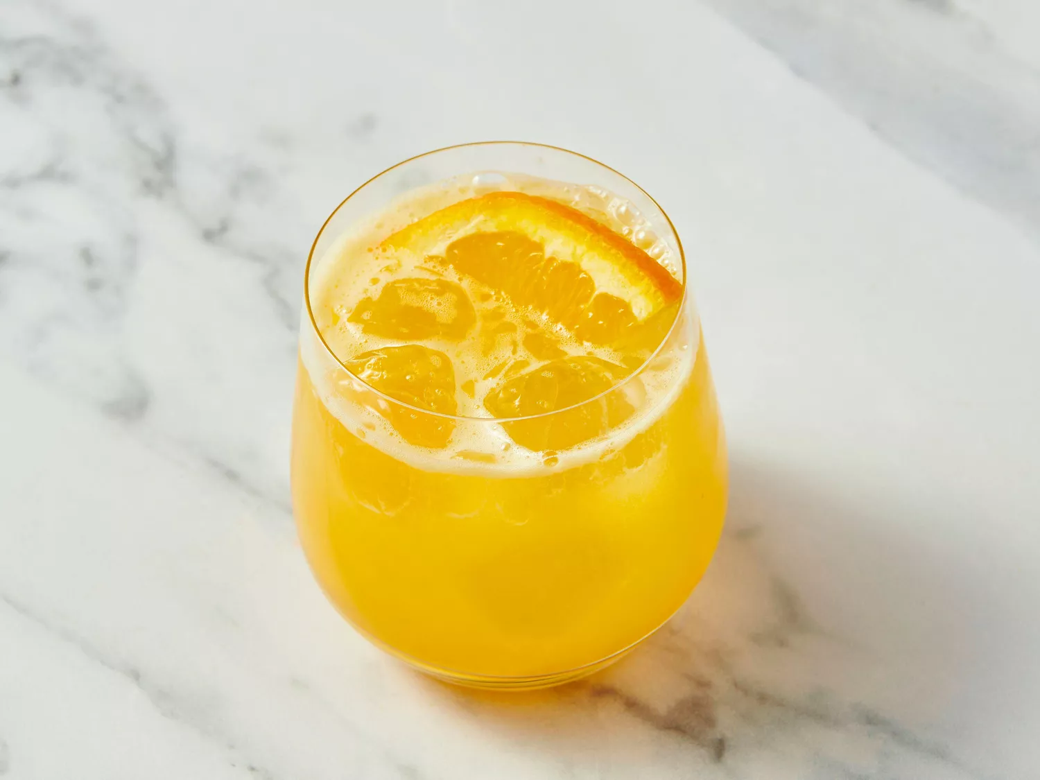 A glass of orange cocktail with ice and a slice of orange, placed on a marble surface