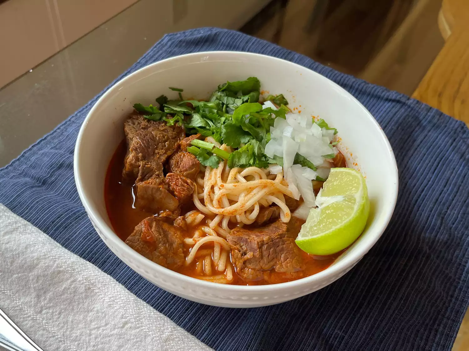 Bowl of ramen in beef birria broth with chunks of beef, cilantro, onion, and lime wedge on blue placemat