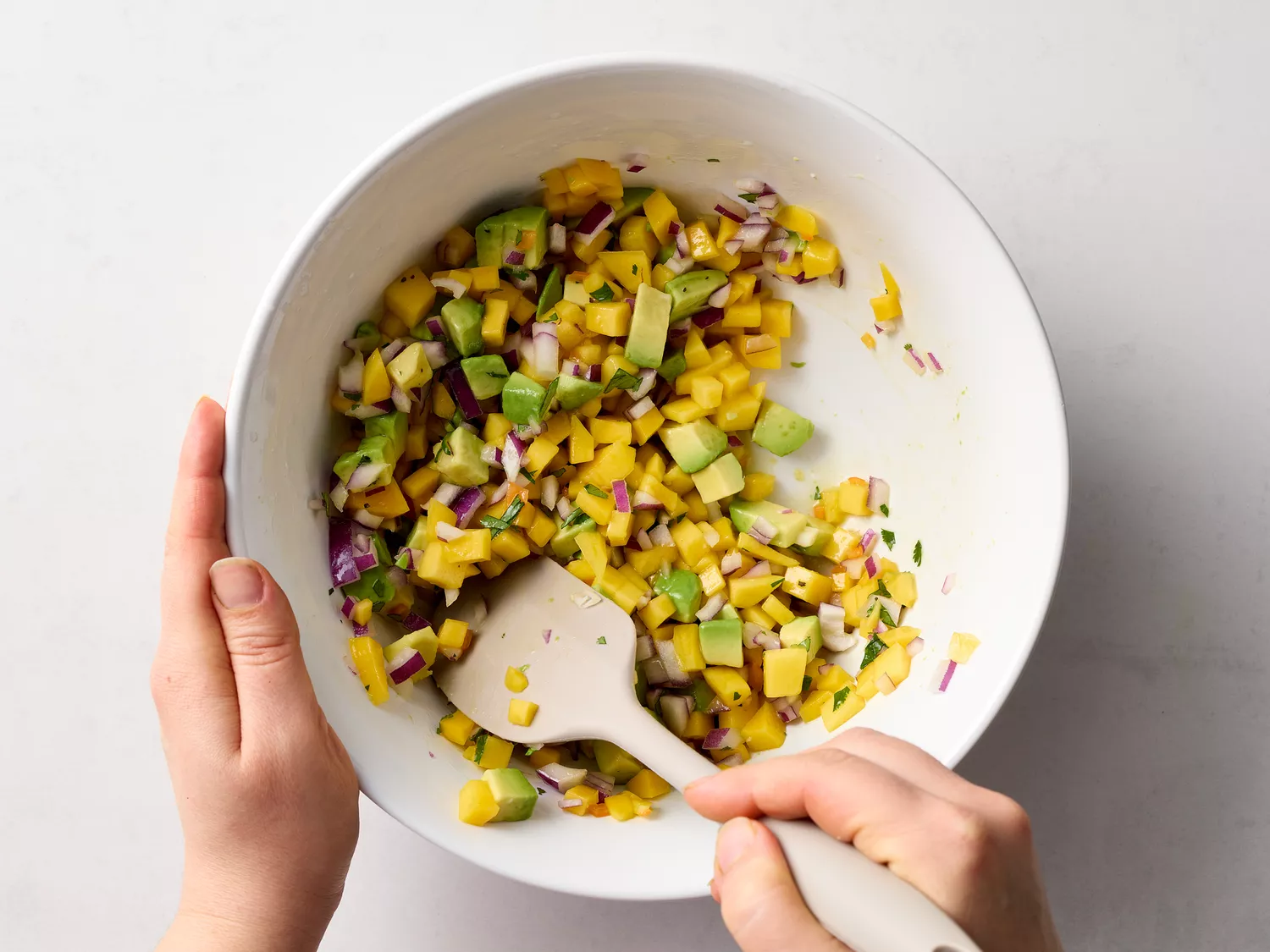 Hands mixing diced mango, avocado, onion, and cilantro in a bowl
