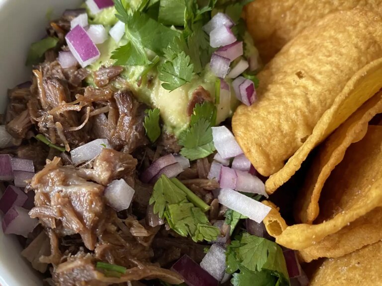 Closeup of carne asada in a bowl garnished with guacamole, chopped red onion, and cilantro with tortilla chips on the side