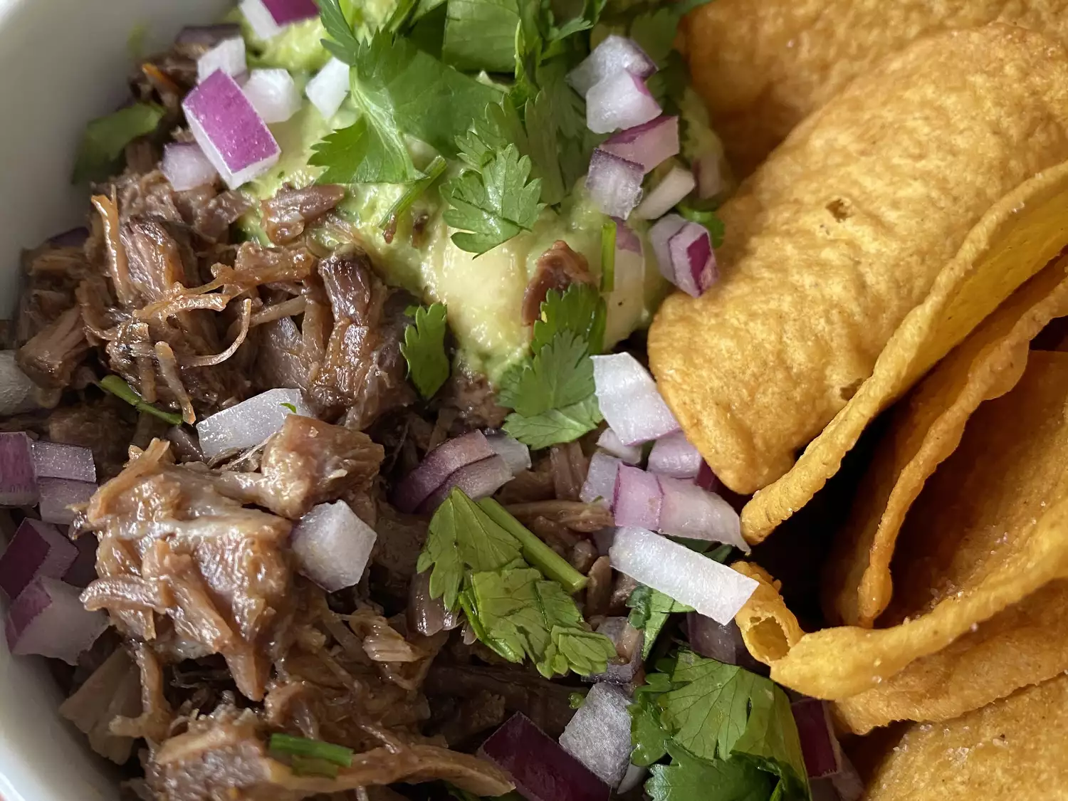 Closeup of carne asada in a bowl garnished with guacamole, chopped red onion, and cilantro with tortilla chips on the side