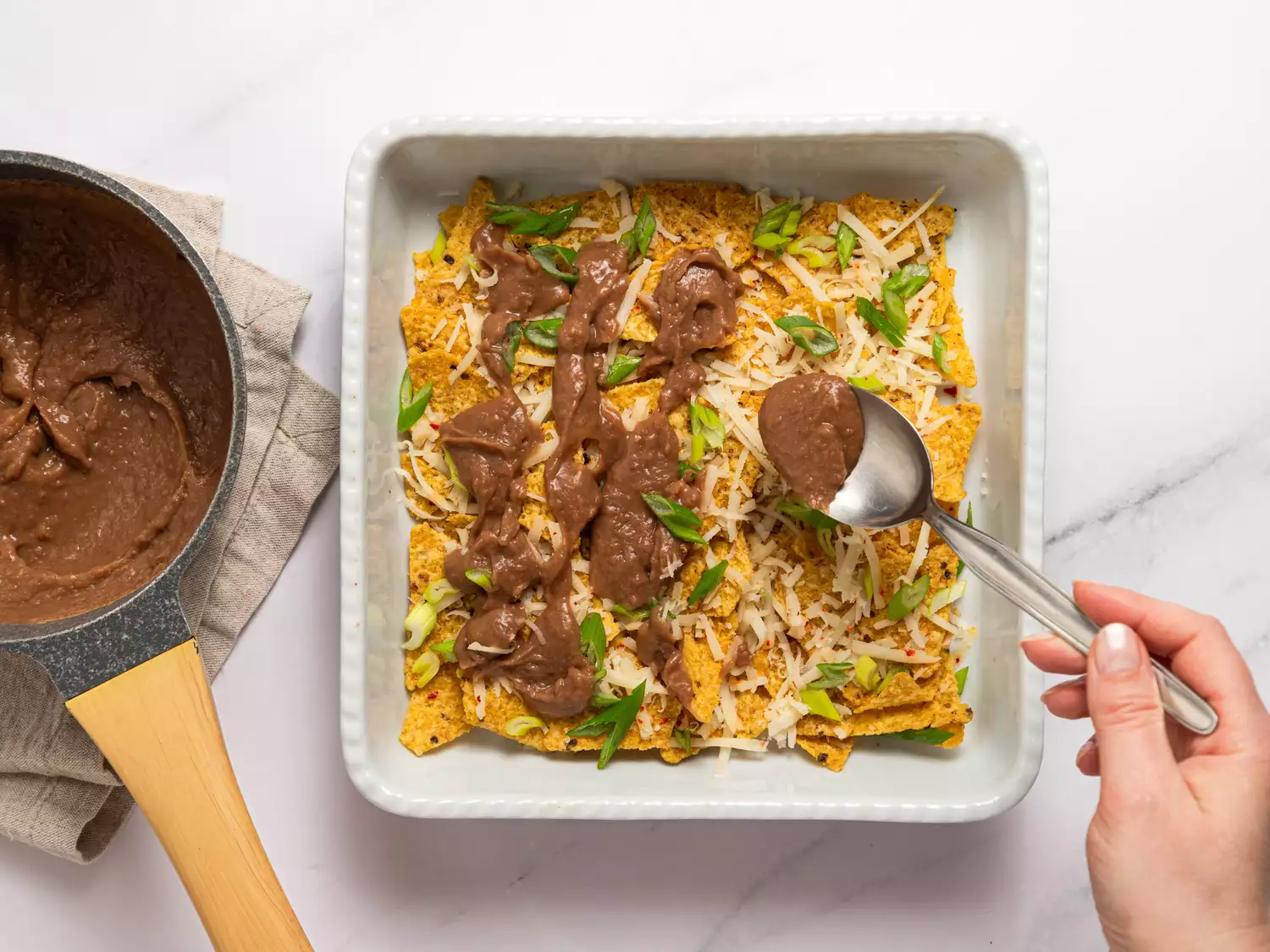 Spoon spreading refried beans over nacho chips in a baking dish.
