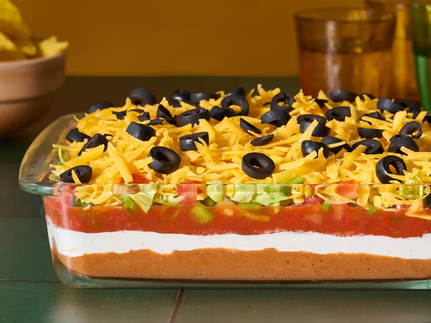 A glass baking dish with seven layer taco dip, topped with shredded cheese and sliced black olives