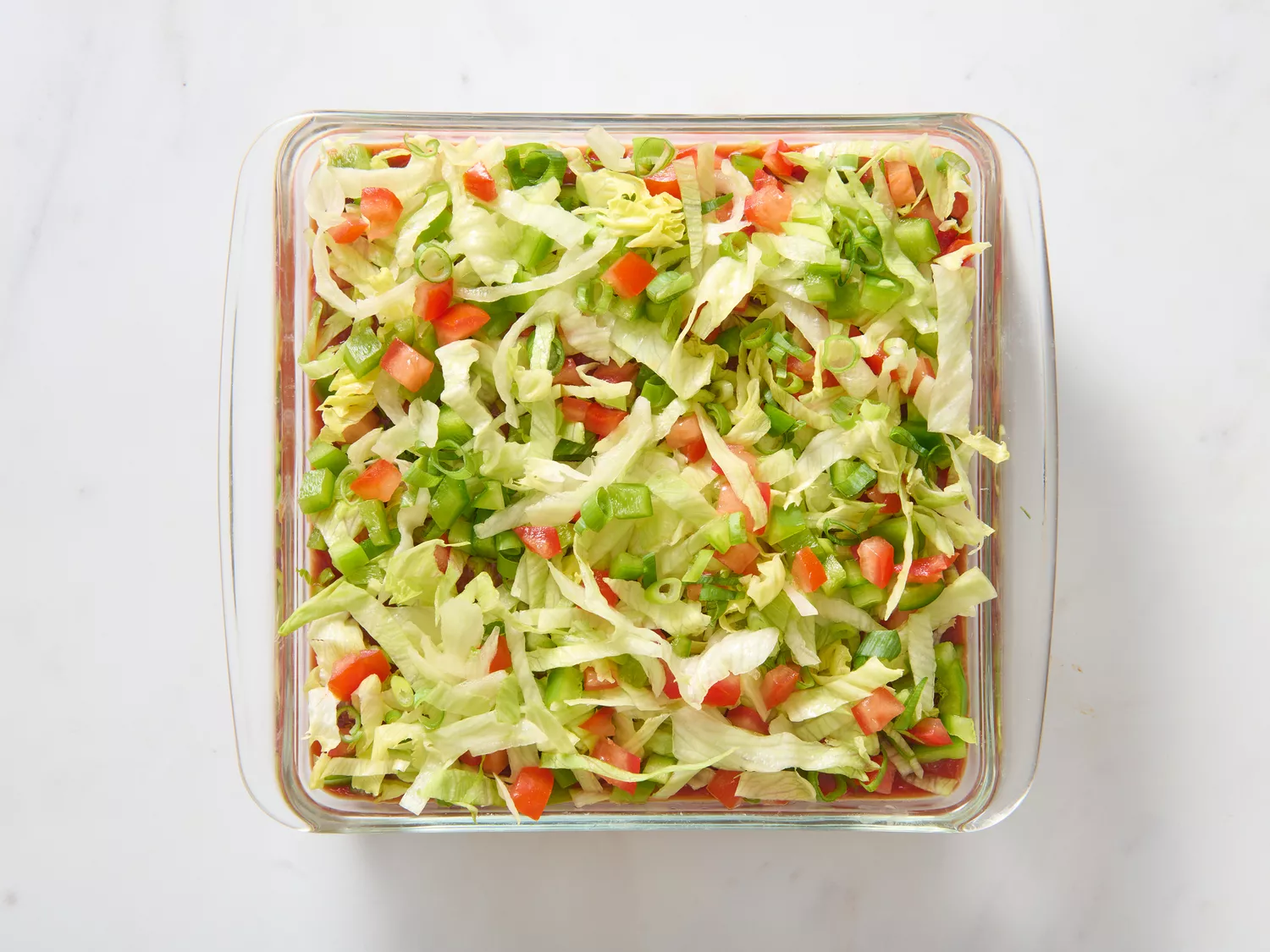 A glass baking dish layered with refried beans, sour cream-cream cheese mixture, salsa, lettuce, tomato, and bell pepper