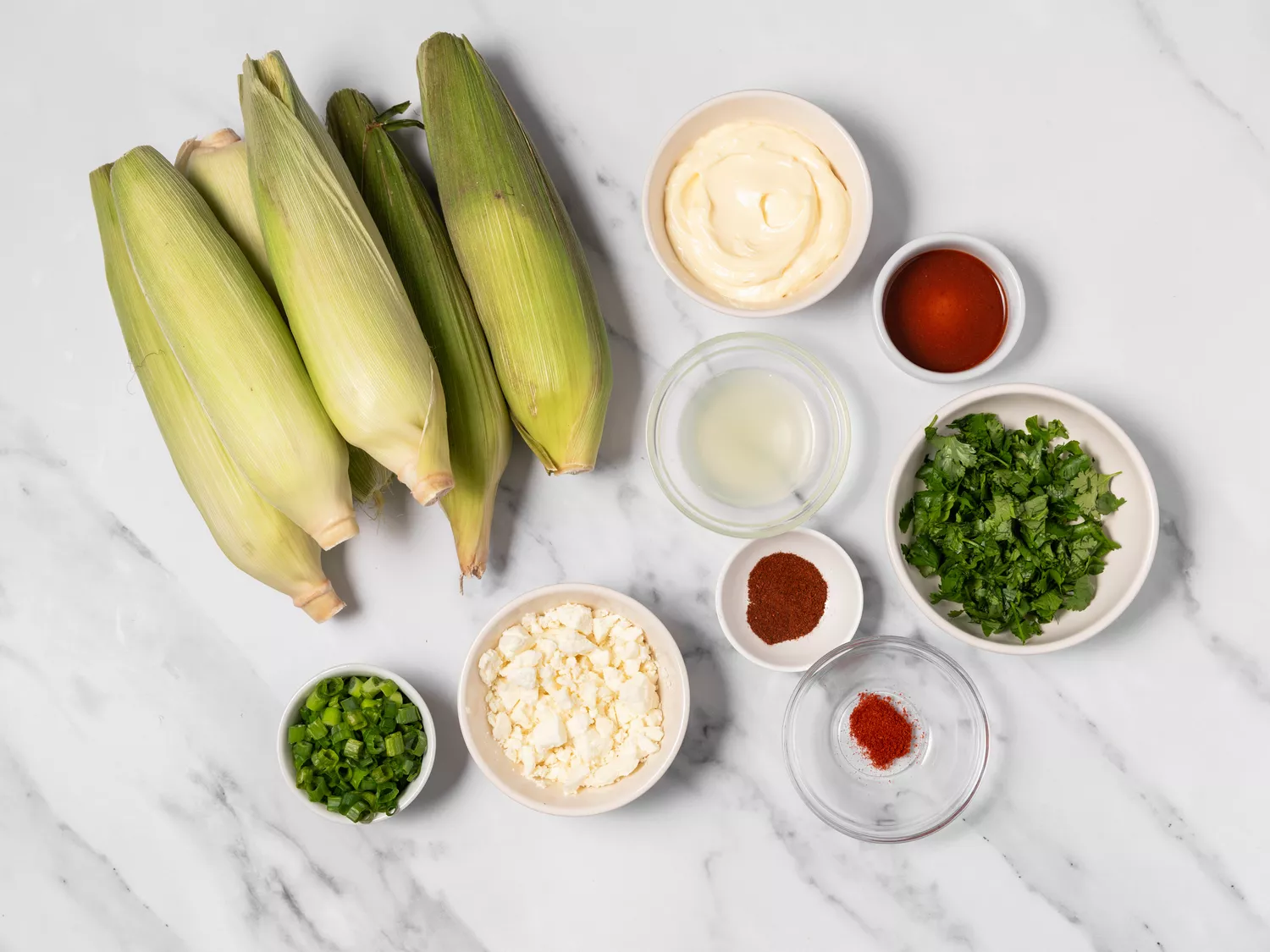 Fresh uncooked ears of corn with various ingredients for elote salad.