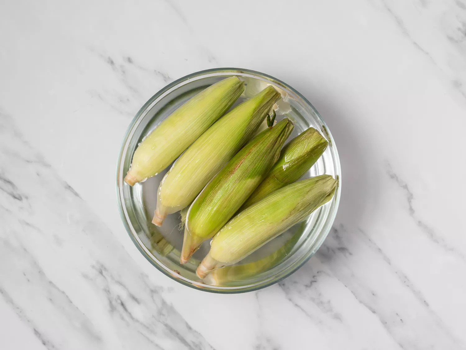Ears of corn soaking in water in a glass bowl.