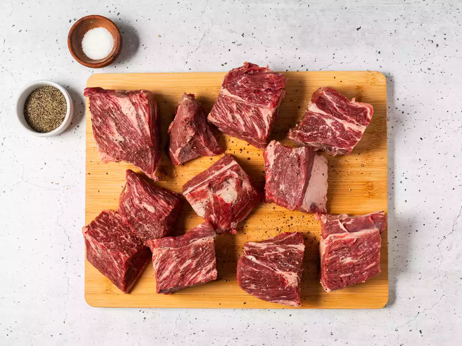A cutting board with cubed beef, topped with salt and pepper