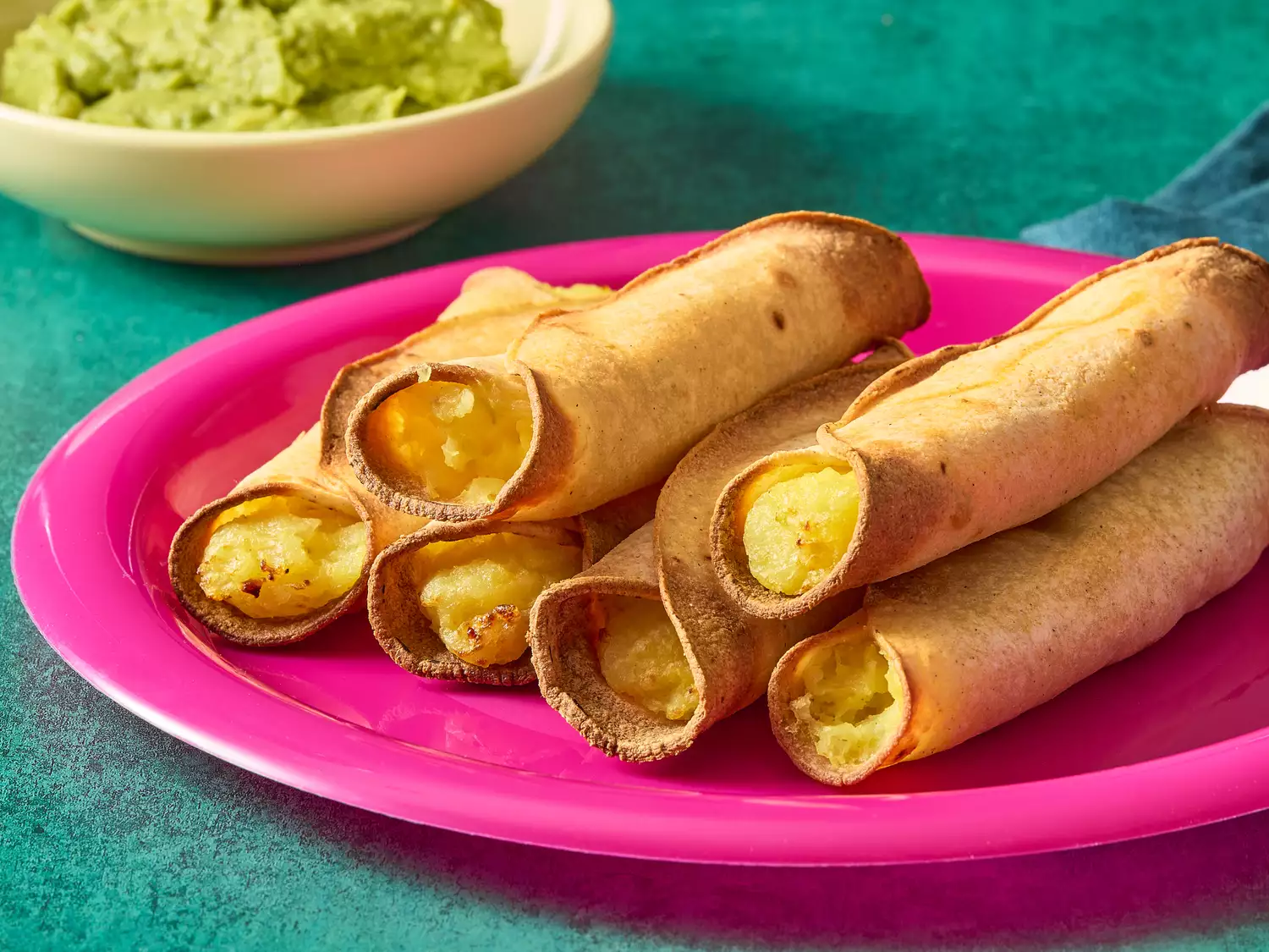 A pink plate with six stacked vegan air fryer taquitos served with a bowl of guacamole