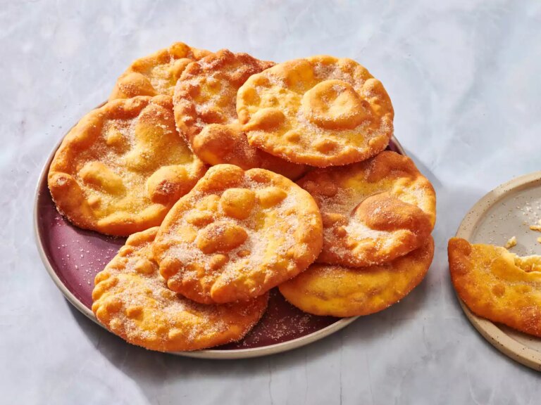 Plate of golden, sugar coated buñuelos on a maroon dish.