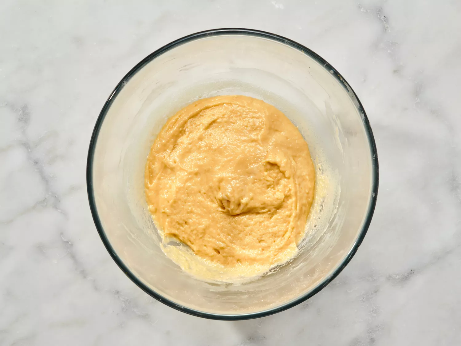 Thick dough mixture in a glass bowl on marble surface.