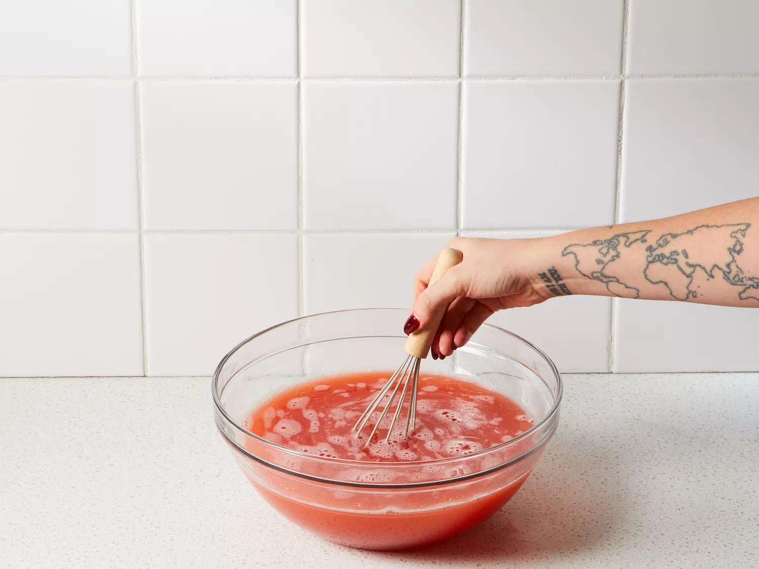 A hand, holding a whisk, whisking together ague de fresa in a large glass bowl