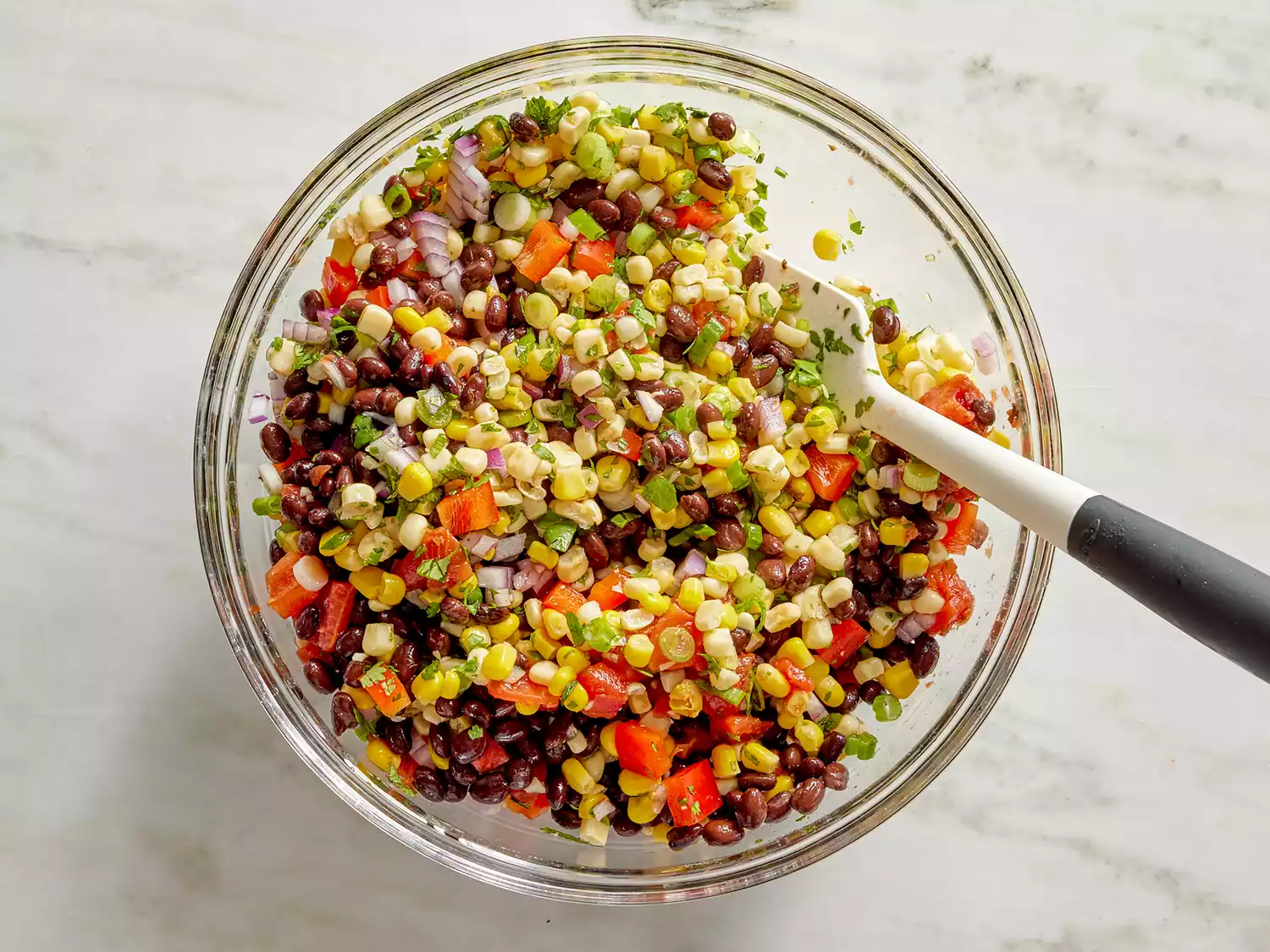 A large bowl with black beans, yellow and white corn, tomatoes, cilantro, green onions, red onion, bell pepper, and garlic being stirred with a spoon