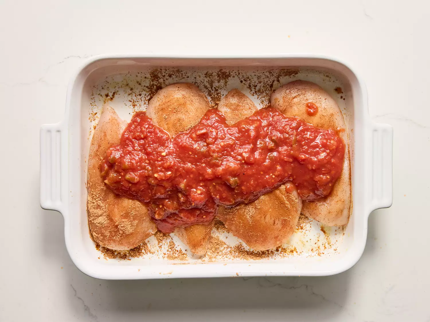 A baking dish with raw chicken breasts topped with seasoning and salsa