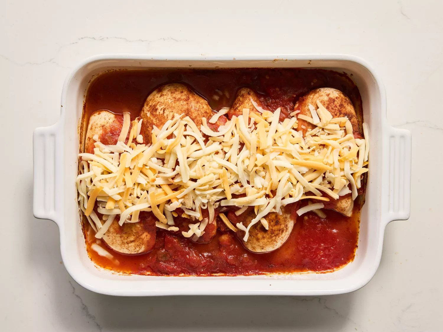 A baking dish of cooked chicken breasts in salsa, topped with shredded cheese