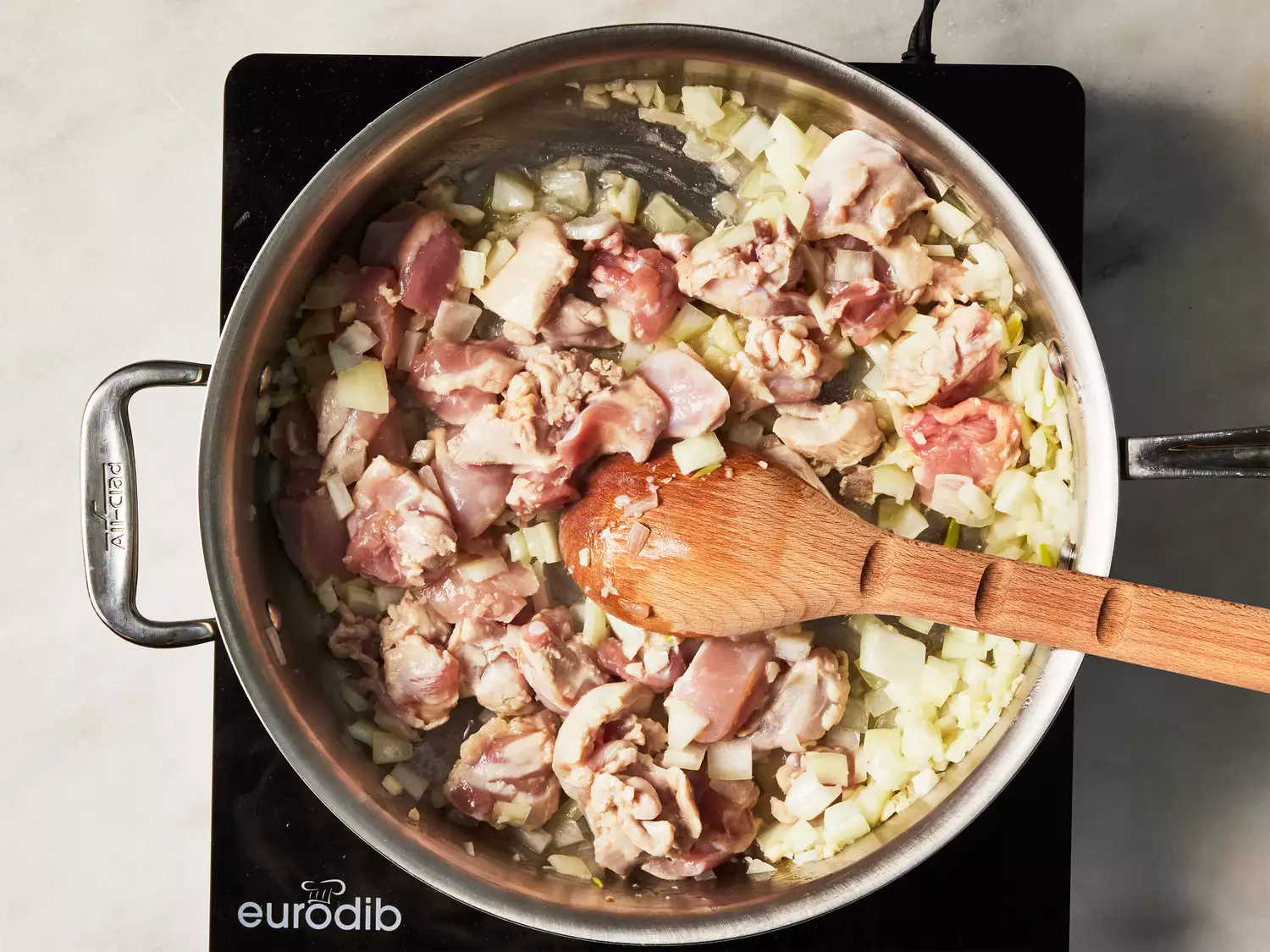 A large pan with pieces of chicken, onion, and garlic being stirred with a wooden spoon