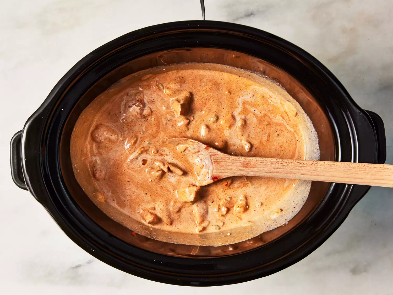 A crock pot with slow cooker butter chicken being stirred with a wooden spoon