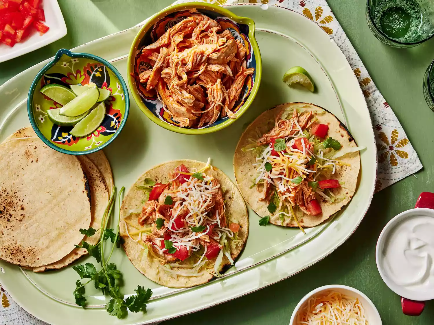 A platter with a bowl of shredded chicken, a bowl of lime wedges, stacked tortillas, and two chicken tacos topped with shredded cheese