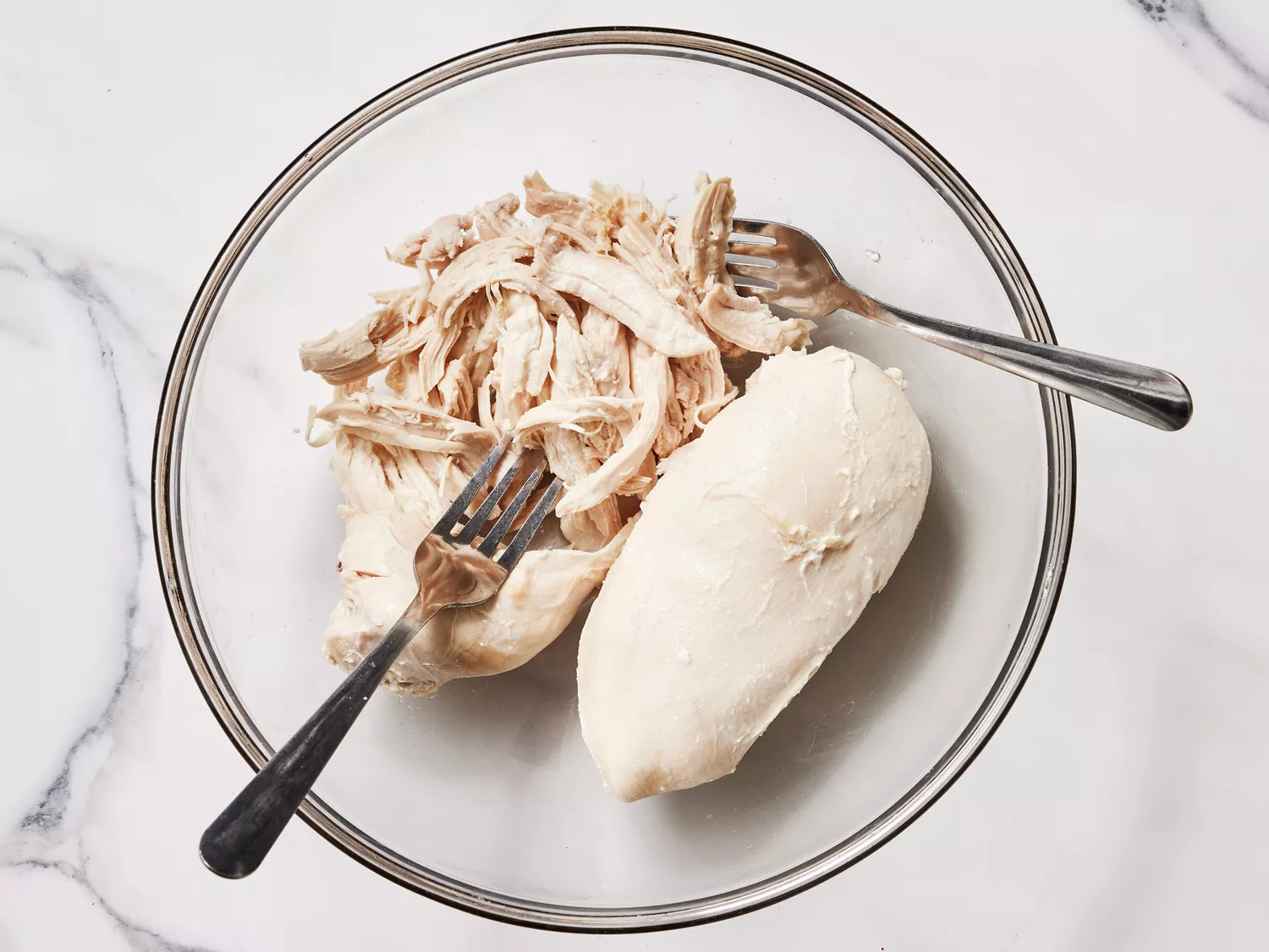 A glass bowl with two boiled chicken breasts being shredded with two forks