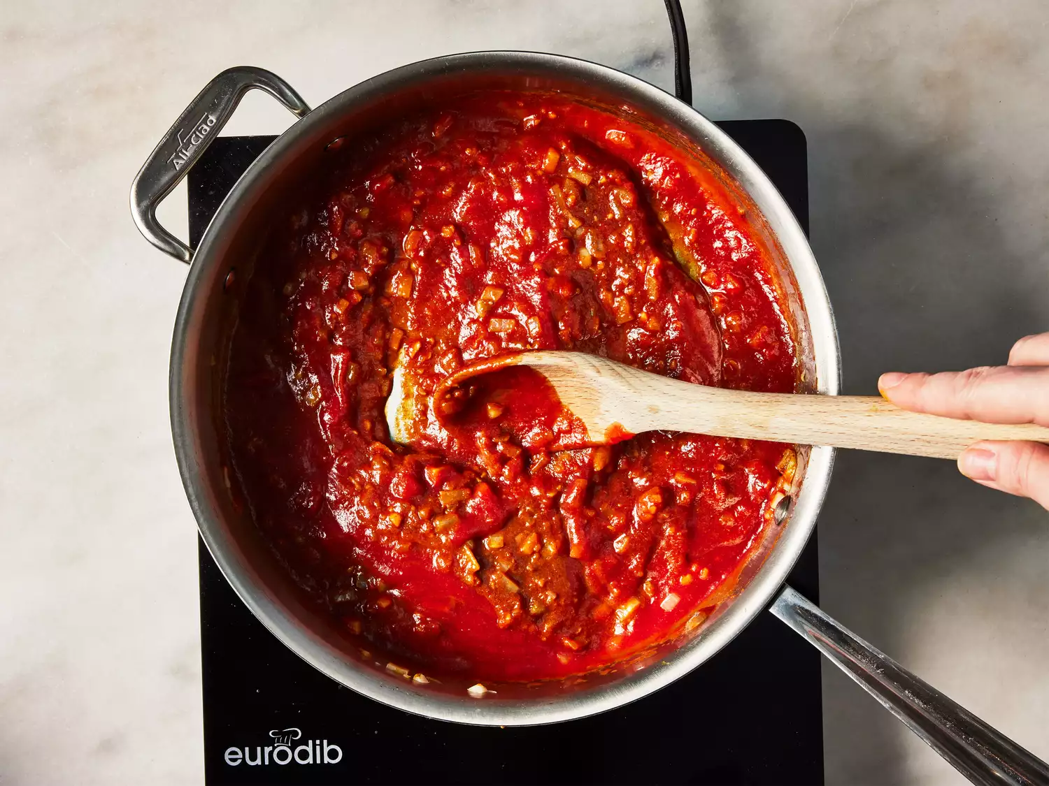 A wooden spoon stirring tomato sauce into a pan of seasoned chopped onions
