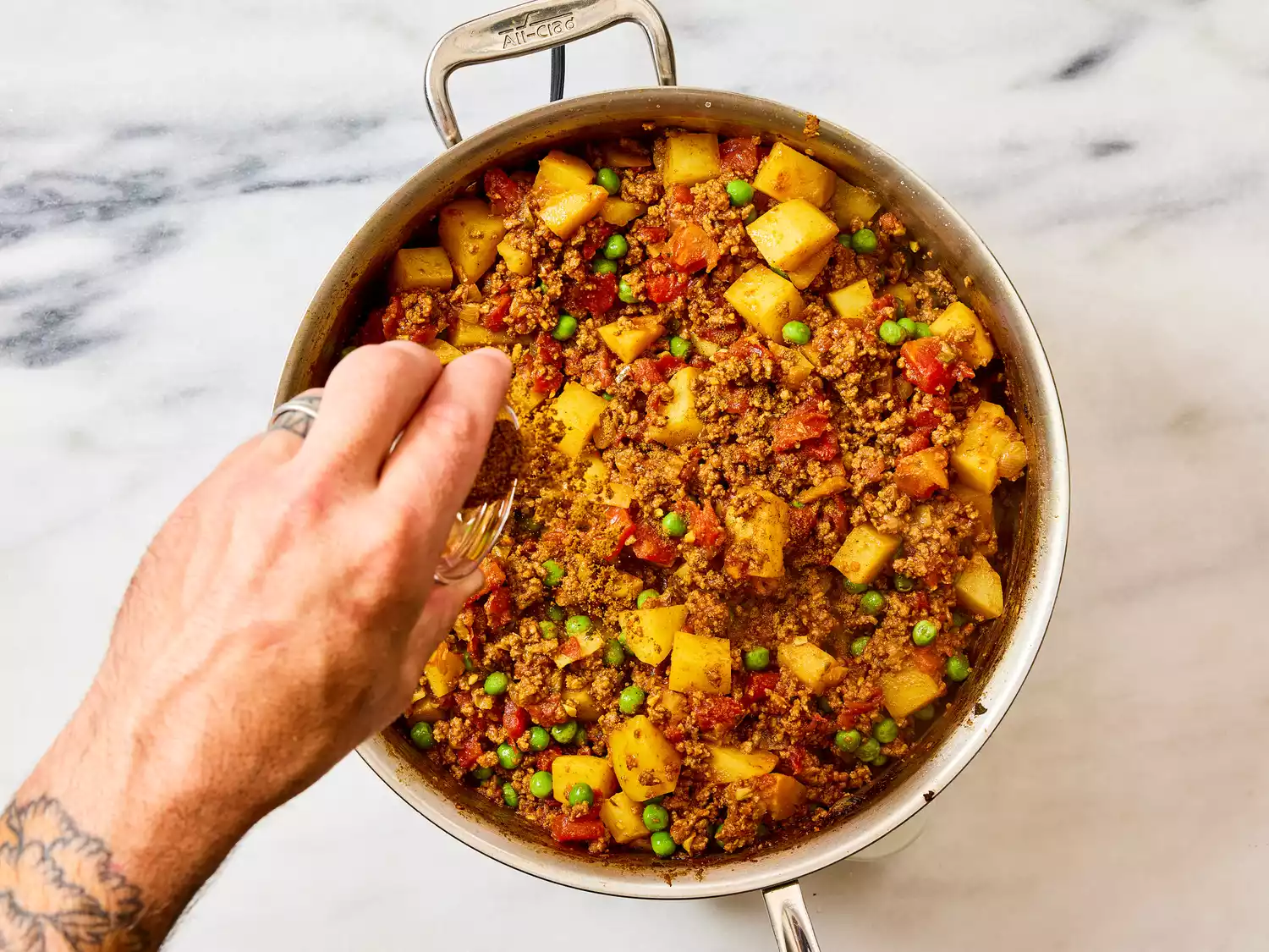 A hand sprinkling garam masala over a pan of keema aloo