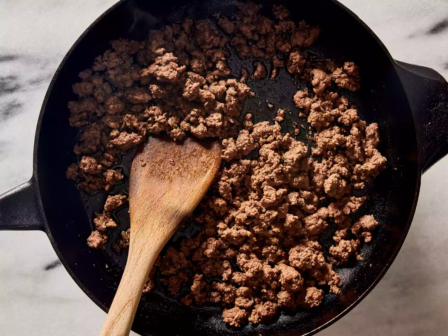 A cast iron skillet with cooked ground beef being stirred with a wooden spoon