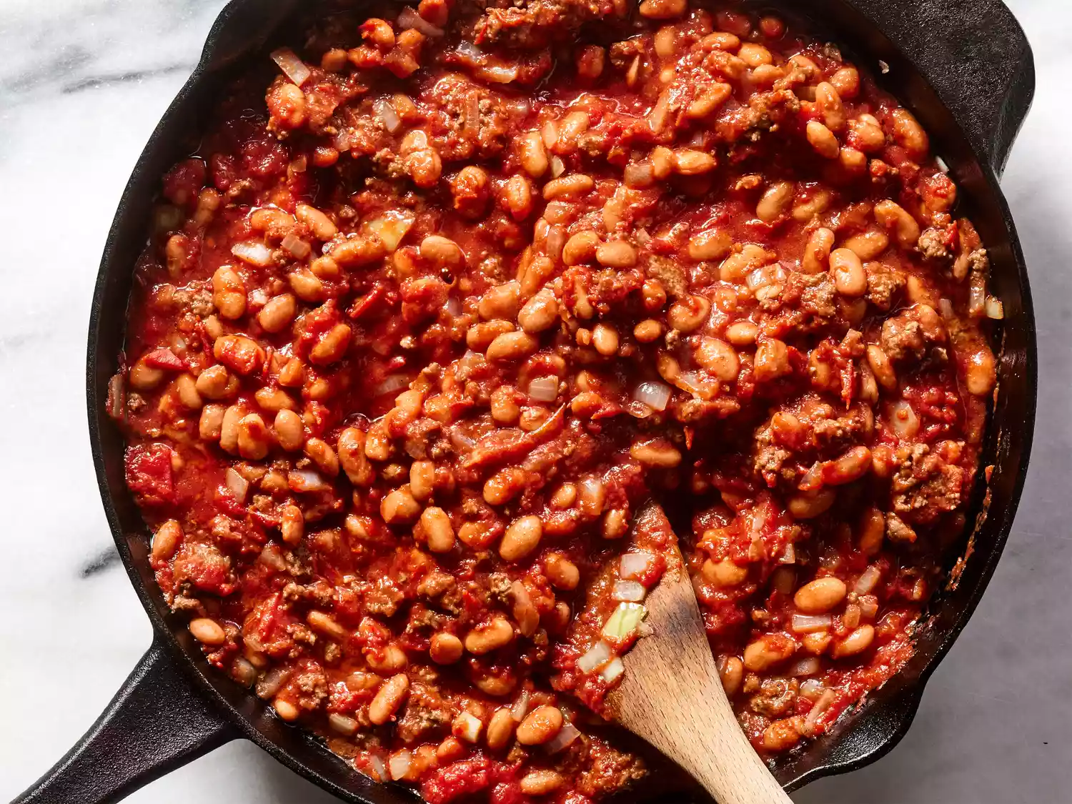 A cast iron skillet with ground beef, chopped onion, and pinto beans bring stirred in tomato sauce