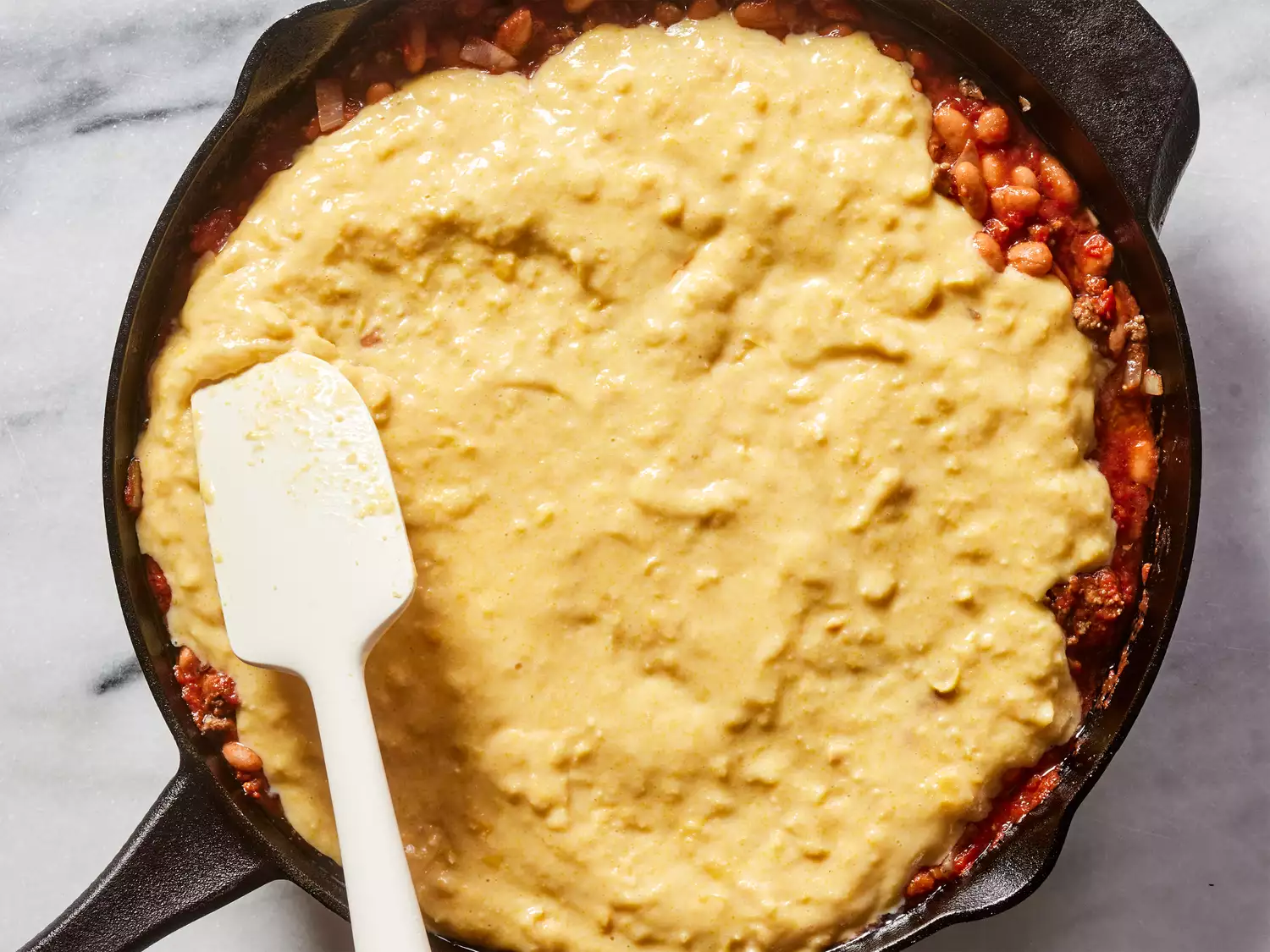 A spatula spreading a cornbread-corn mixture over the skillet with ground beef