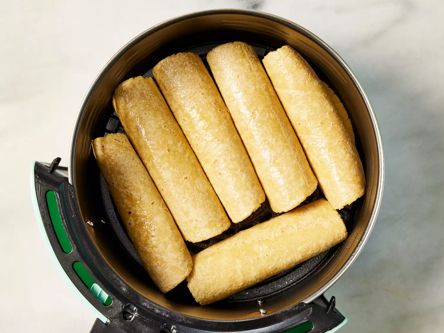An air fryer basket with six rolled up chicken taquitos