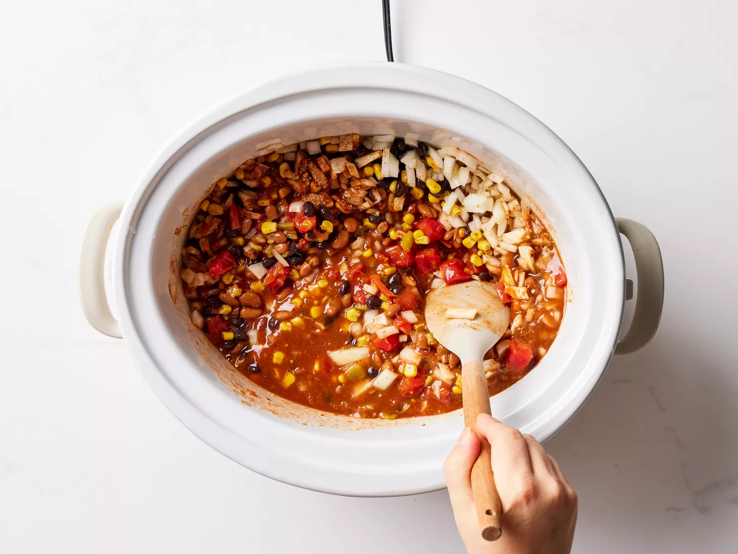 A slow cooker with diced tomatoes, chili beans, black beans, corn, beer, tomato sauce, and onion being stirred with a spatula
