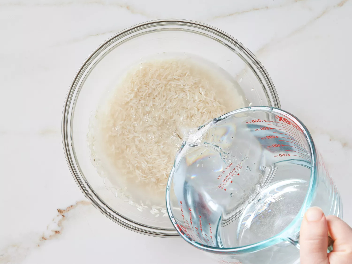 A hand pouring a measuring cup of water over basmati rice in a glass bowl