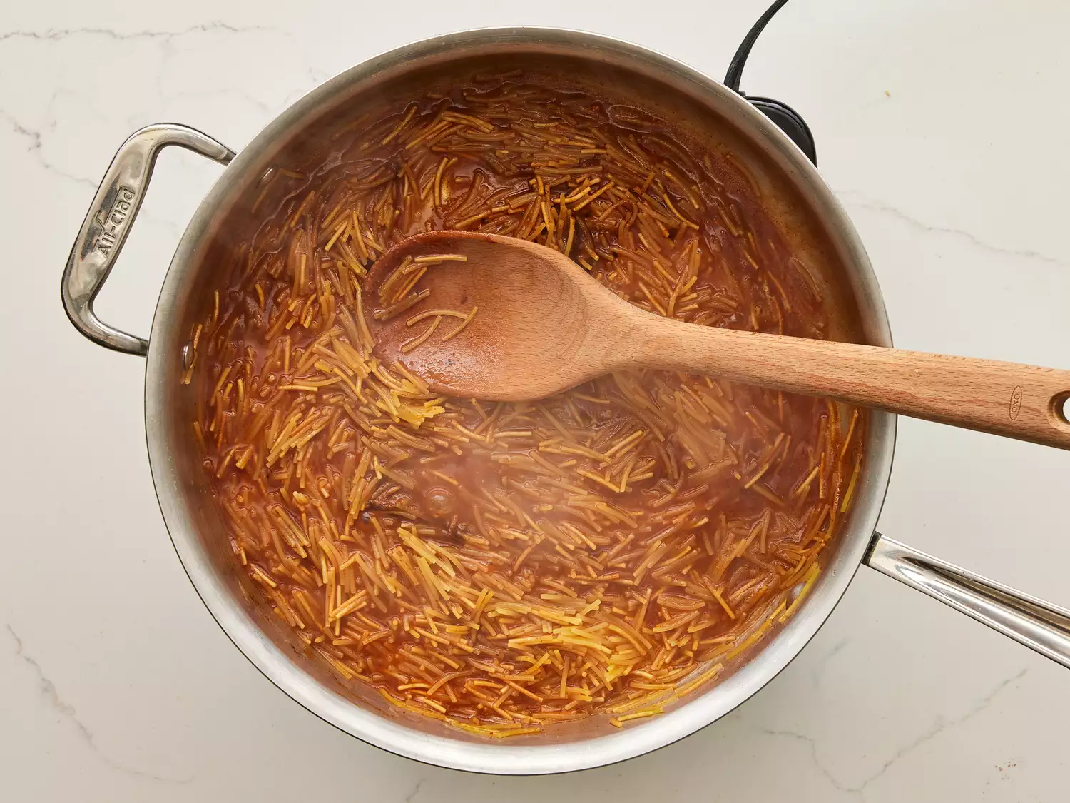 A pot of Fideo pasta in seasoned tomato sauce and water, being stirred with a wooden spoon
