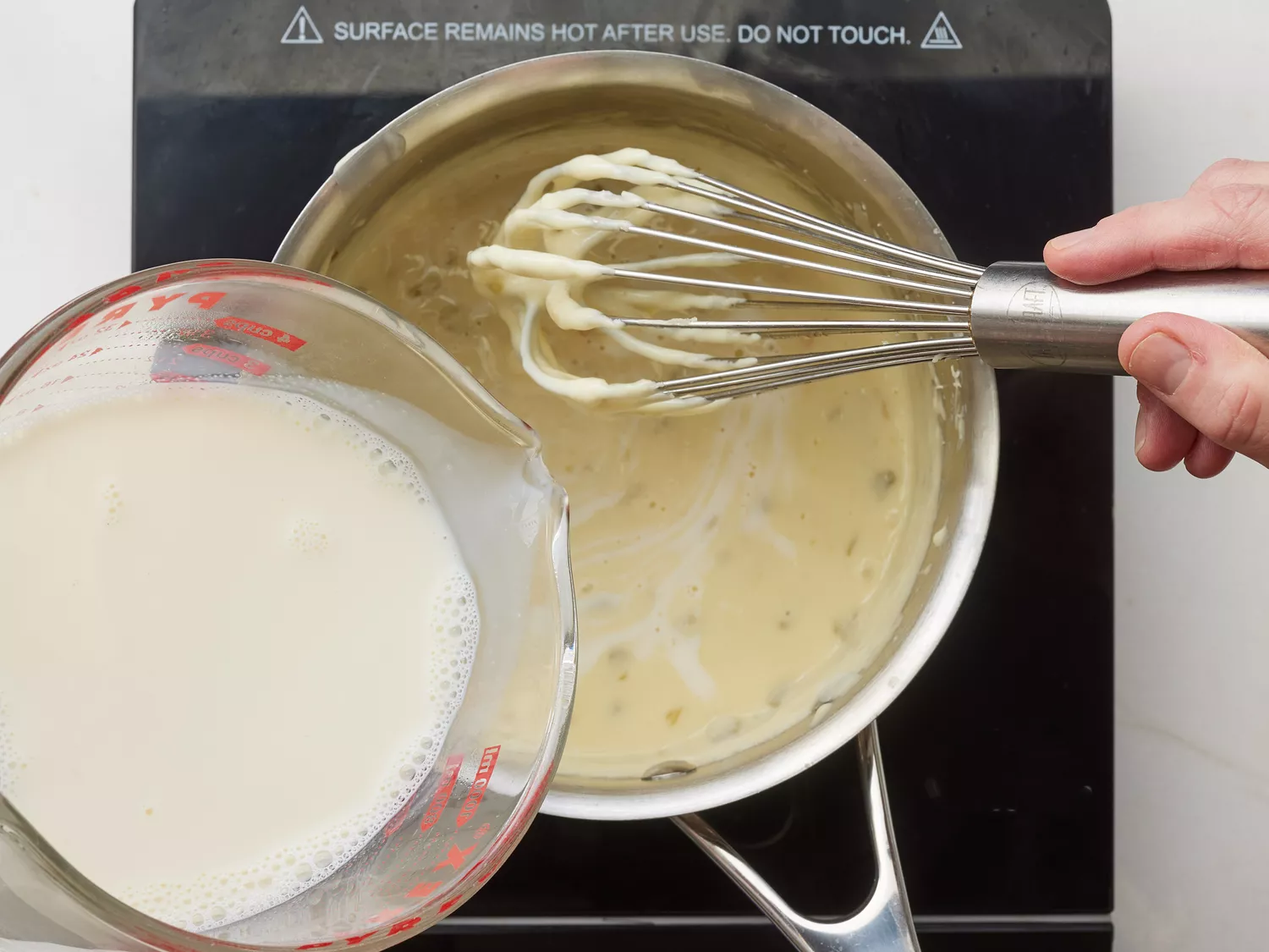 A hand pouring a measuring cup of milk into a saucepan of Mexican queso sauce