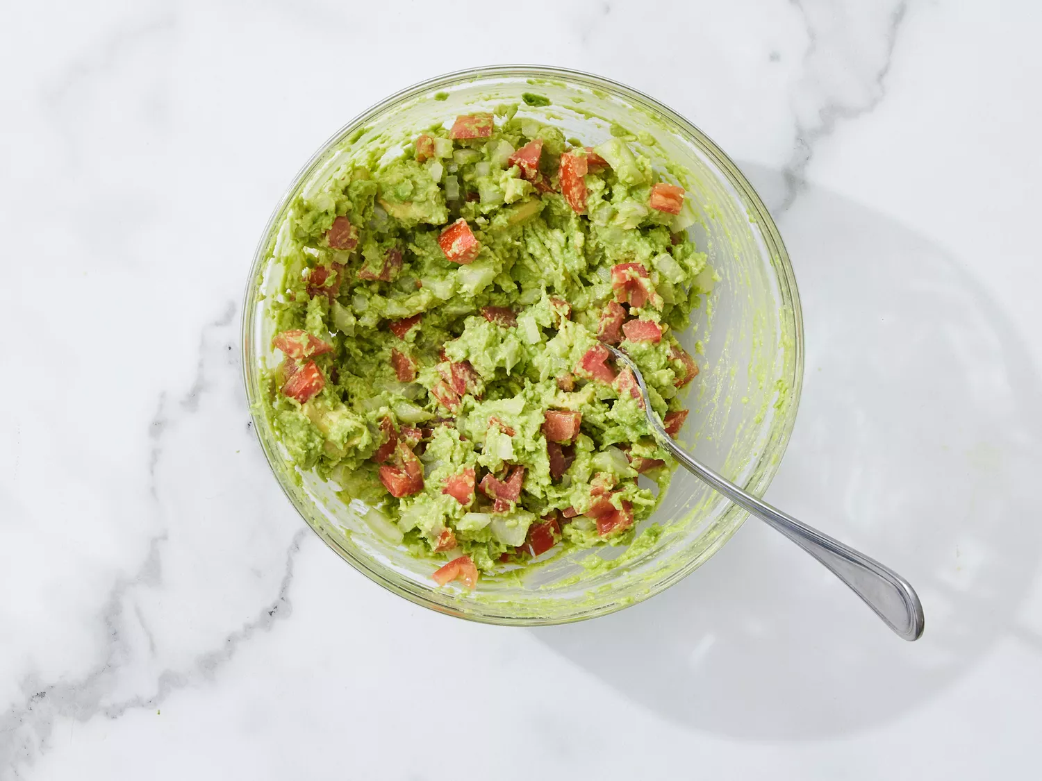 A glass bowl with mashed avocado, chopped tomato, onion, and garlic with a fork