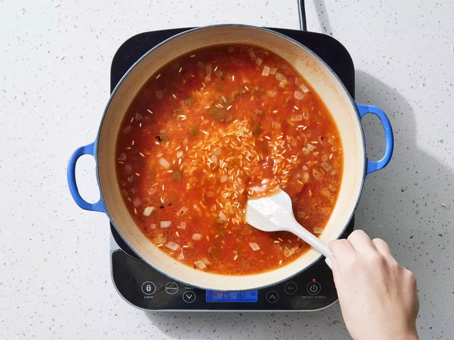 A spatula stirring chicken broth and picante sauce into a large pot of rice