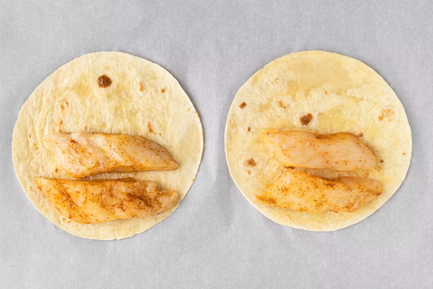 Two tortilla shells with pieces of seasoned fish on a parchment line baking sheet. 