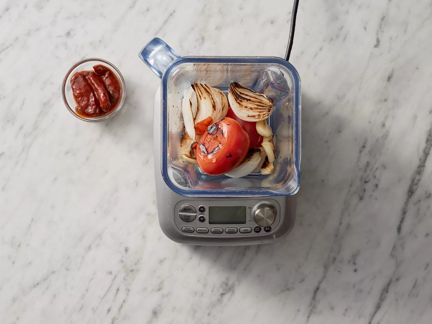 Overhead of cooked vegetables in a blender with chipotle peppers off to the side. 